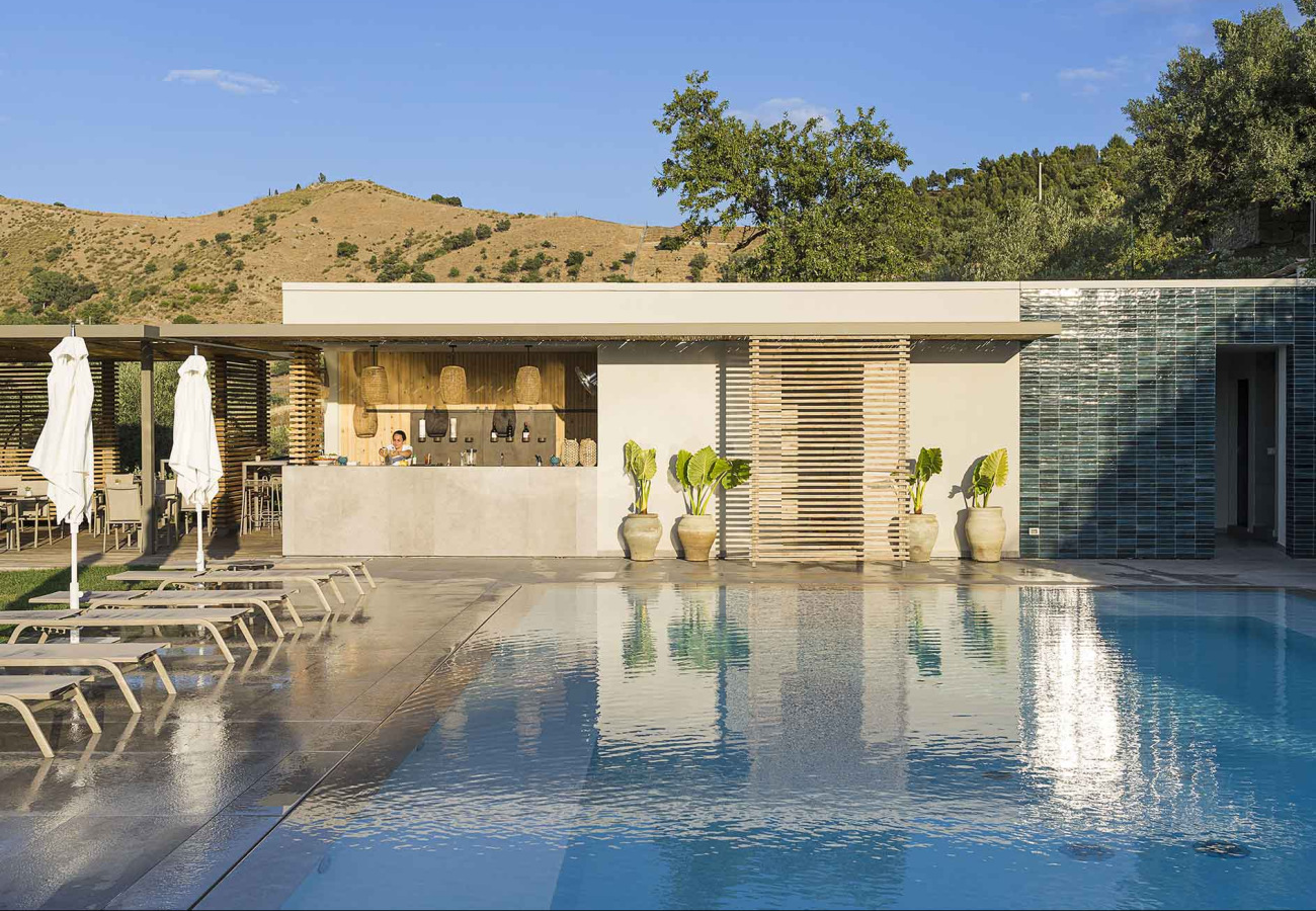 Piscina con bar moderno e lettini, circondata da colline e vegetazione.