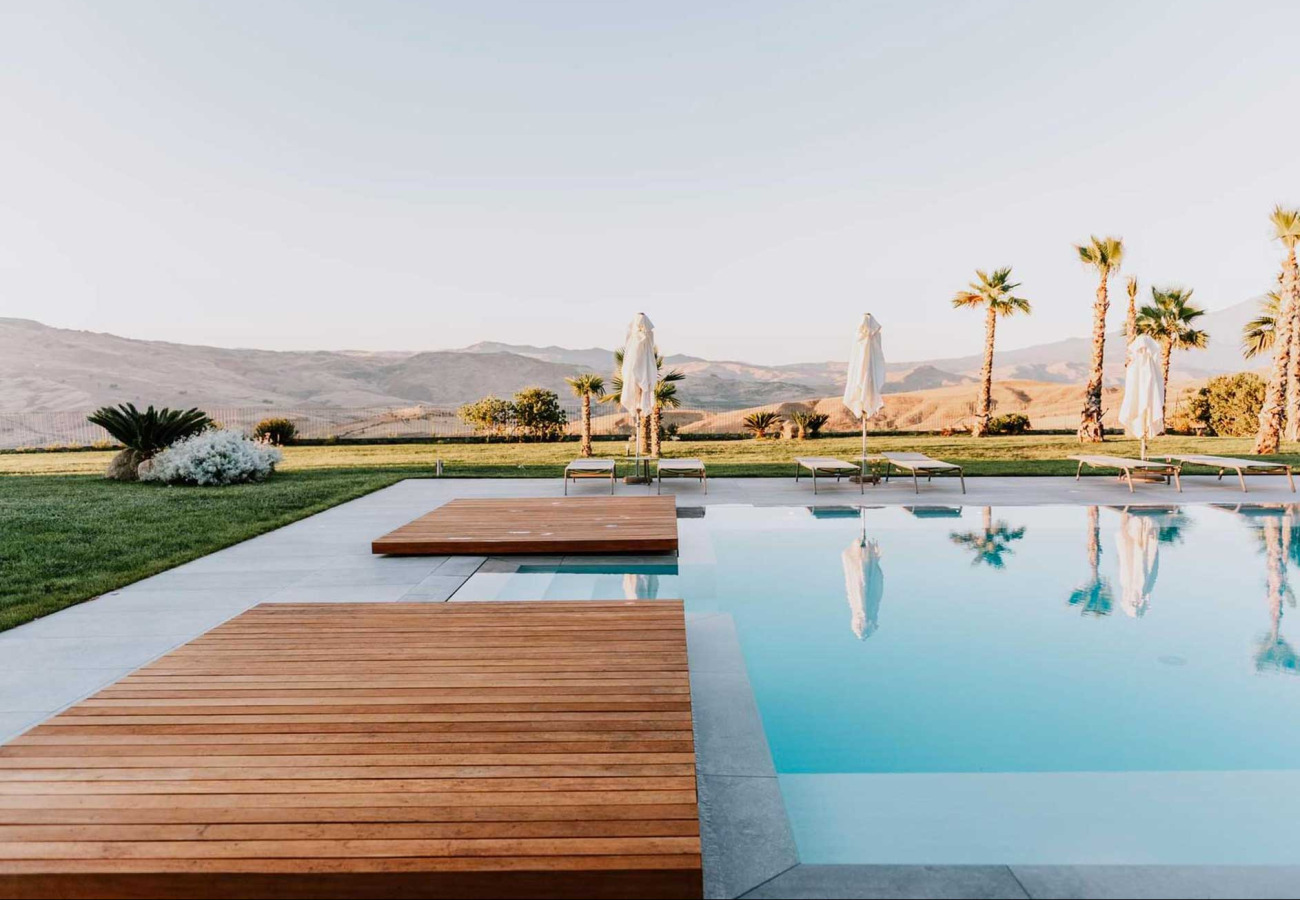 Piscina moderna con vista su colline, palme e lettini con ombrelloni bianchi.