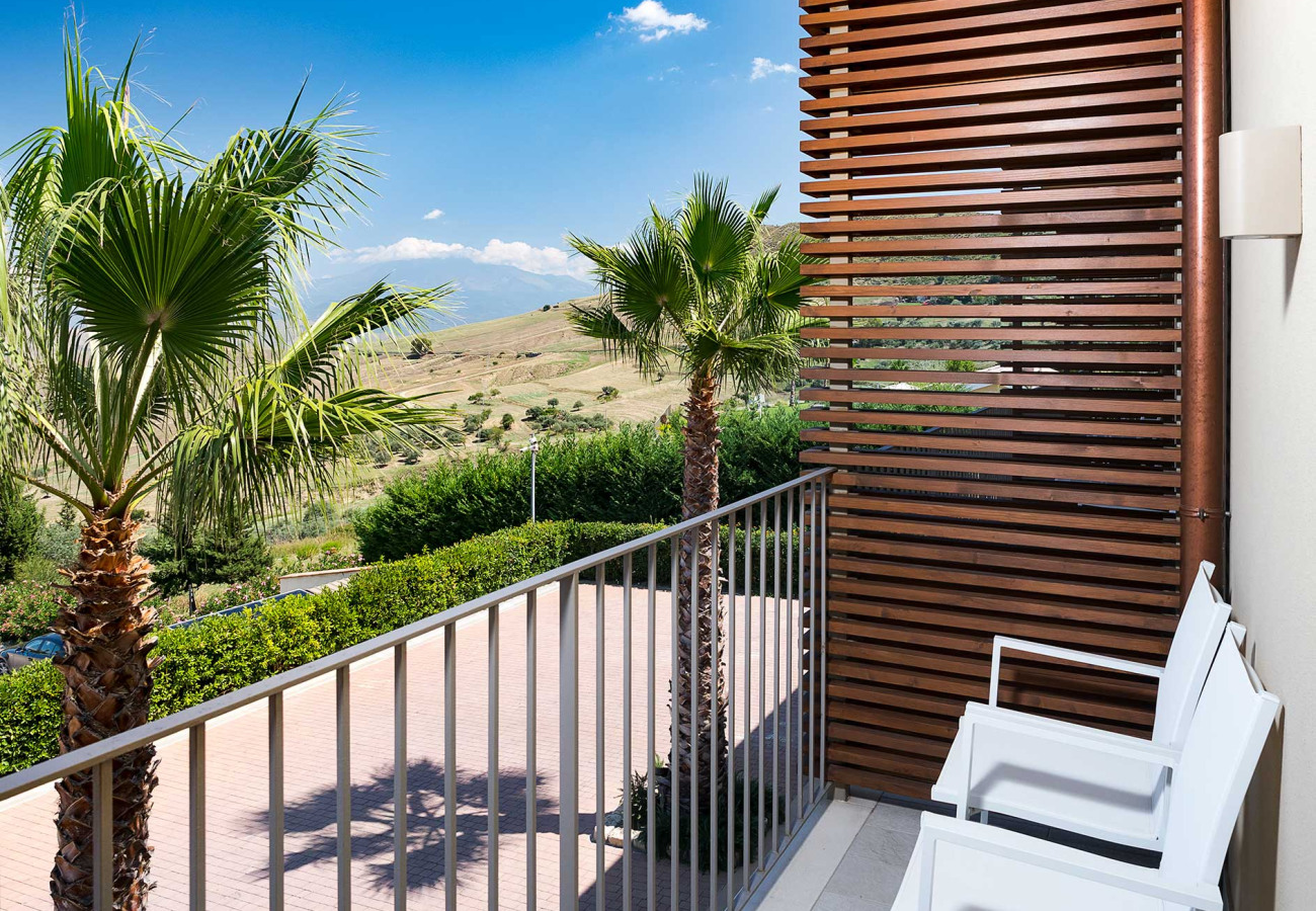 Balcone con sedie bianche, palme e vista su colline verdi sotto un cielo azzurro.