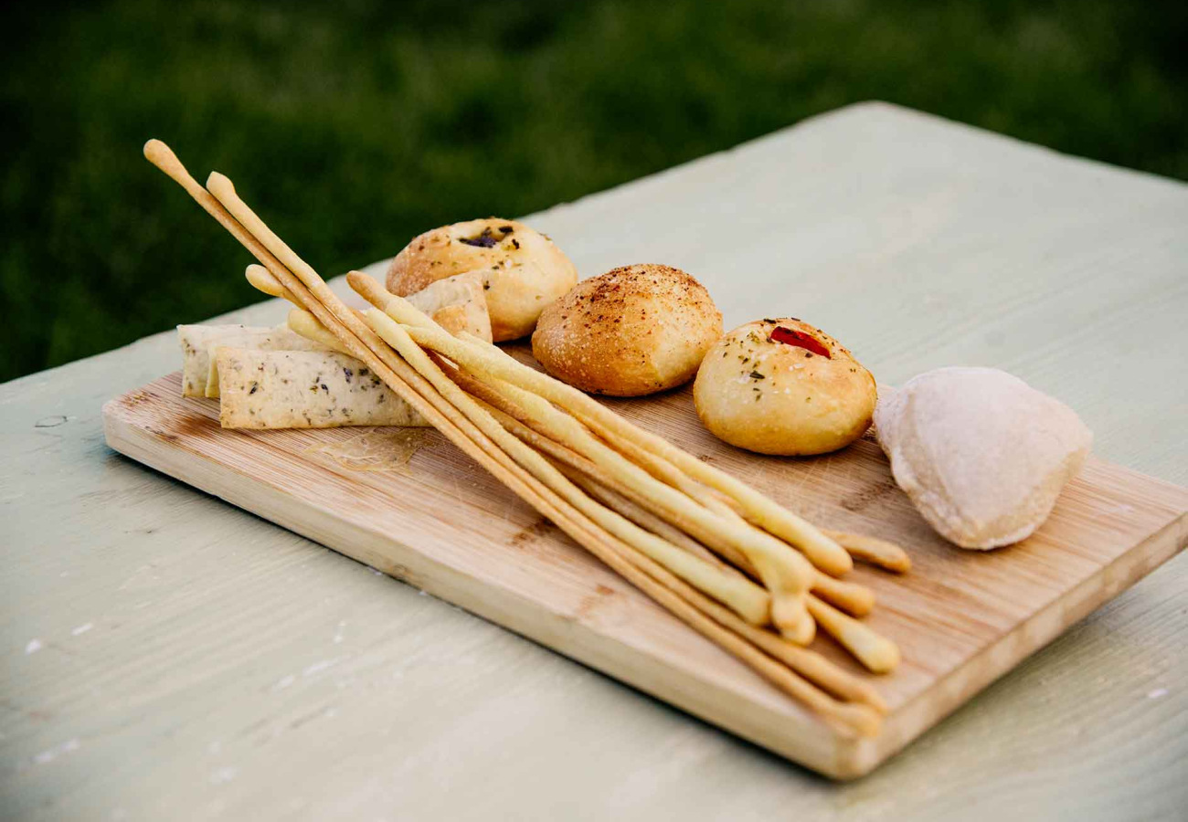 Tagliere con grissini, formaggio e panini assortiti su tavolo di legno.