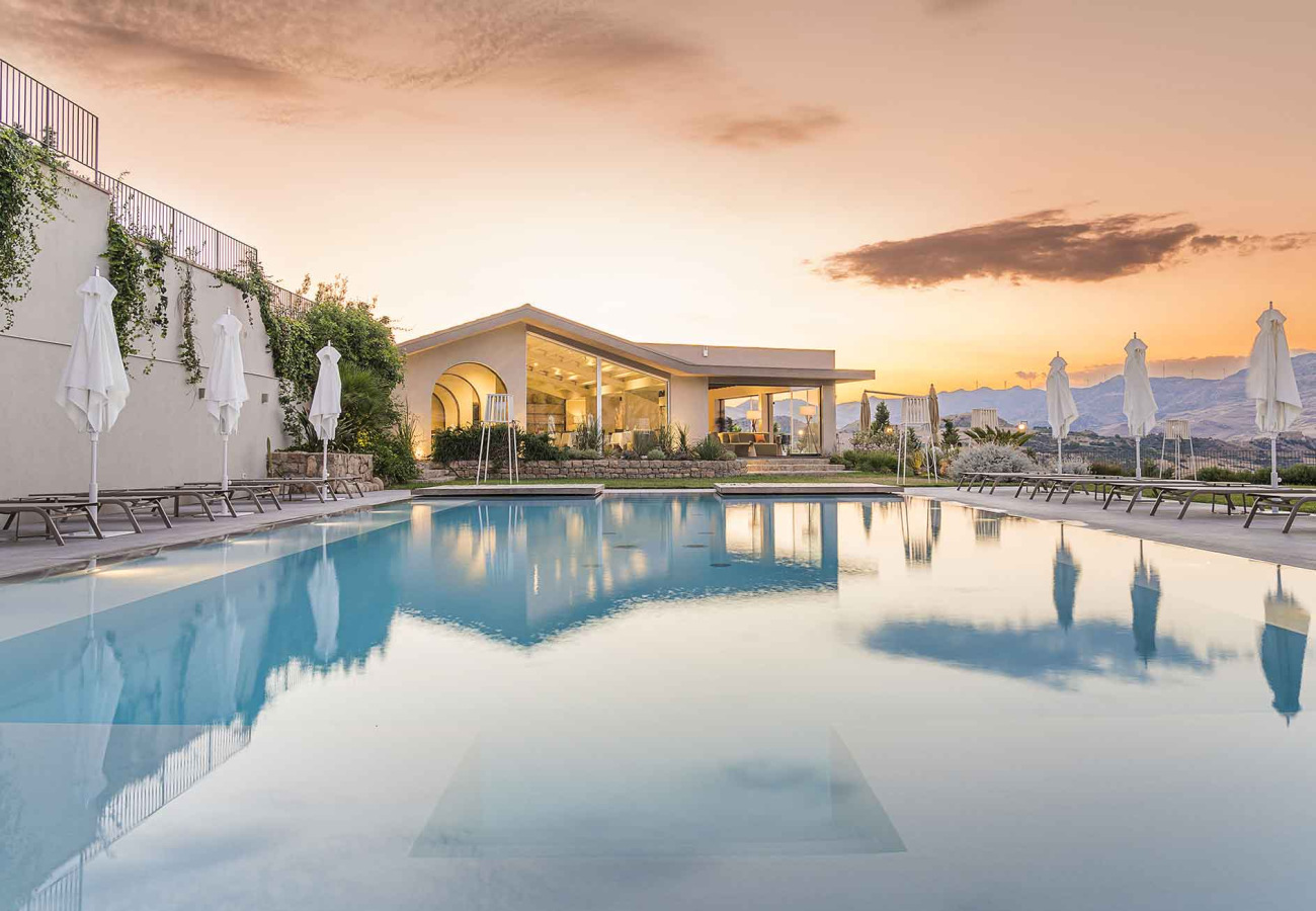 Piscina con vista al tramonto, ombrelloni chiusi e edificio moderno sullo sfondo.