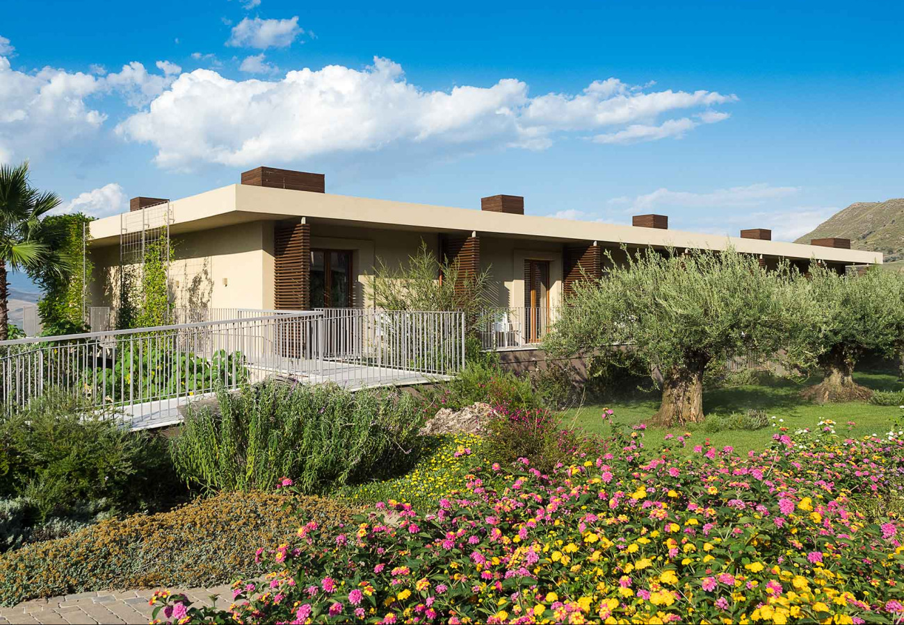 Casa moderna con giardino fiorito e ulivi sotto un cielo azzurro.