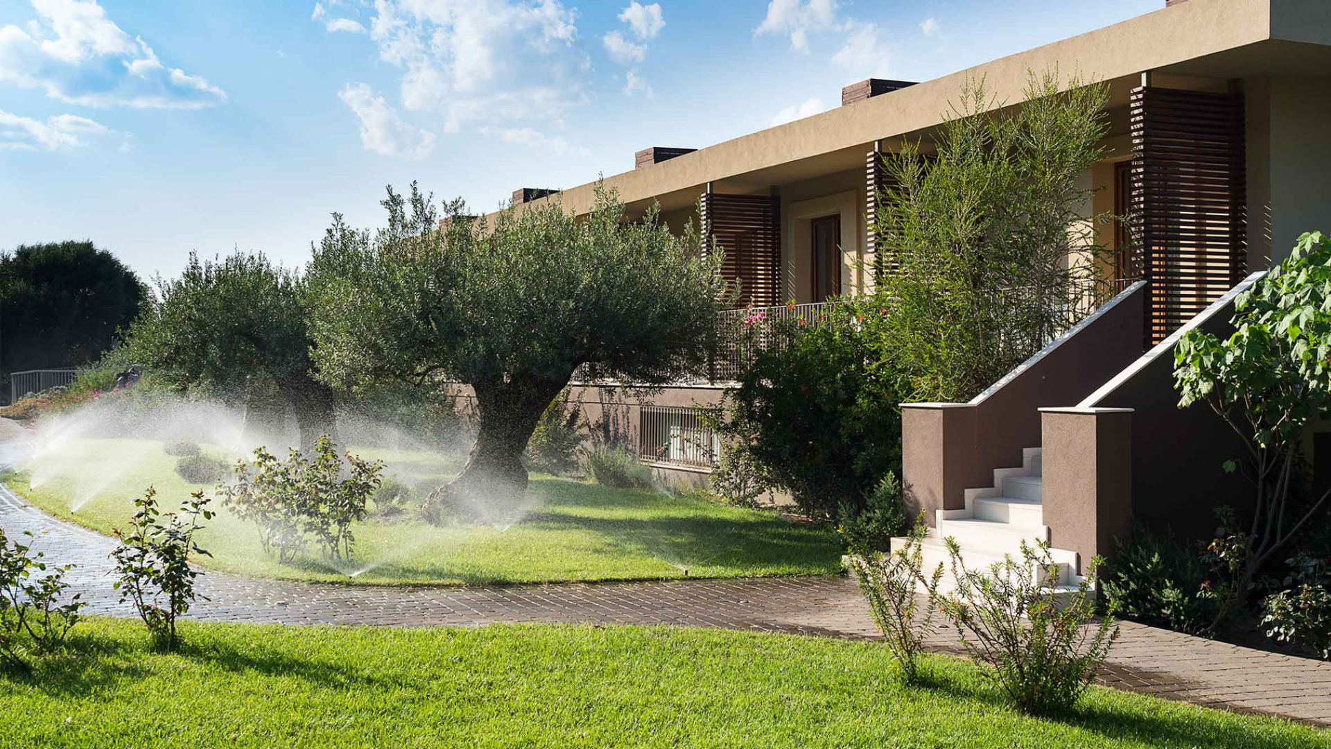 Giardino curato con irrigazione, alberi e un moderno edificio residenziale.